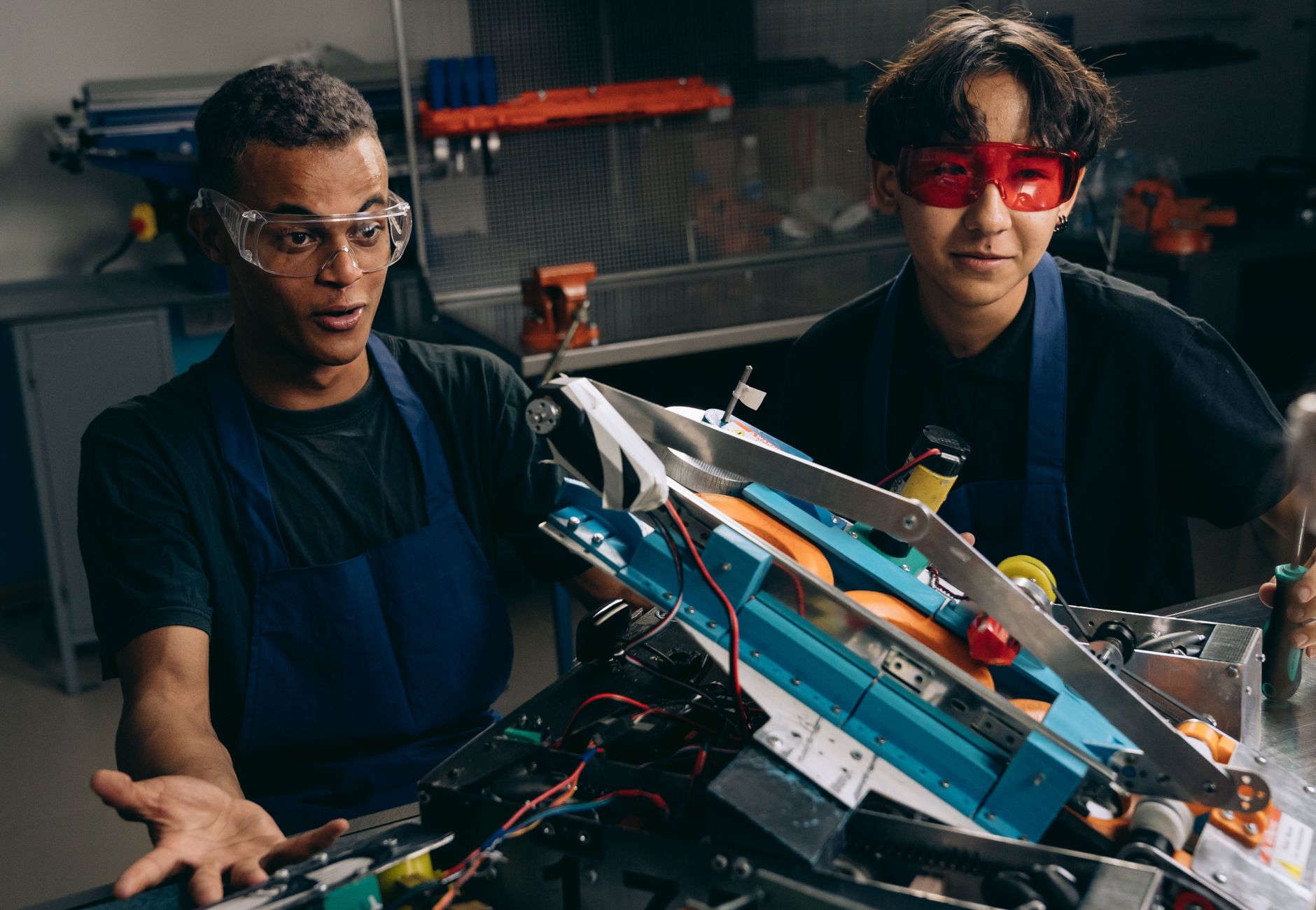 Two men wearing goggles and aprons collaborating on a robotic project in a workshop.