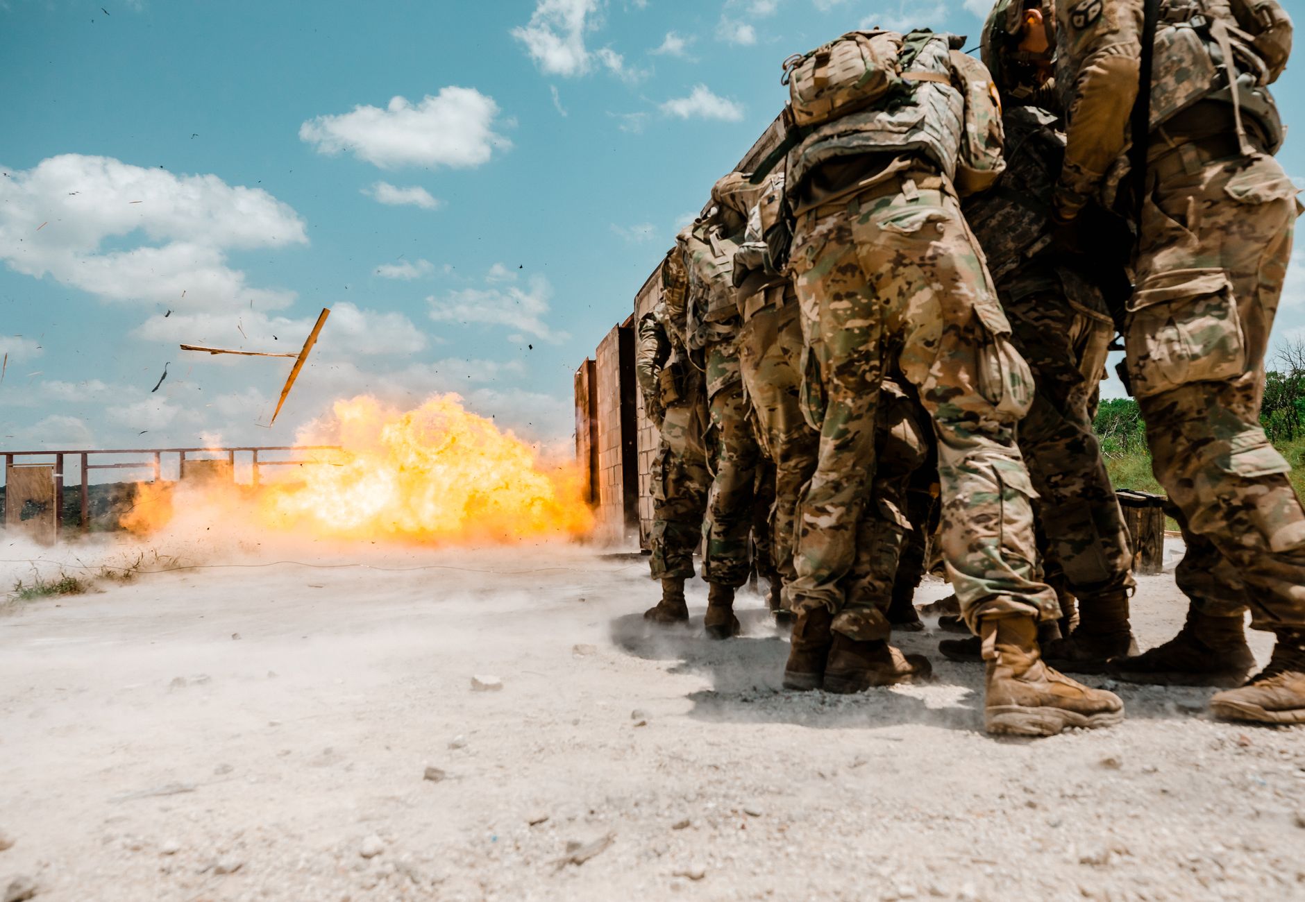 Group of soldiers in military gear facing explosion during training exercise outdoors.